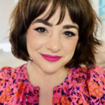 Natania Barron smiles at the camera. She has a bob haircut with bangs, soft brown eyes framed with black eyeliner, and wears a bright pink lipstick that matches her pink and orange print blouse.